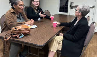 Image of three women sitting at a table having a conversation while two hold red cups and a bag and plate sits in front of the third