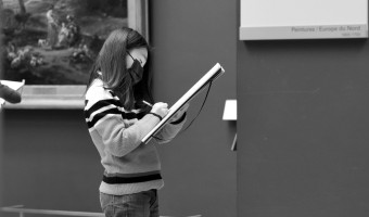 Black and white image of a girl writing on a notepad with a painting behind her and museum label in the foreground by Tommy on Unsplash.      