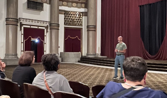 Image of a man in a t-shirt and jeans standing in front of a stage with elaborate columns and a torn red curtain behind him with the backs of people's heads in the foreground as they listen.
