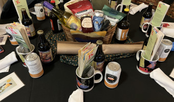 Image of centerpiece with wine, snacks, a hat, and more on a piece of sunflower fabric with a mug, bottle, can, and pamphlets at each table setting from the 2024 Setting the Tables at the KMA Conference in Fort Scott.