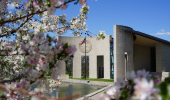 A an exterior photo of the Dole Institute (curved portico with an American flag) and blooming flowers in the foreground.