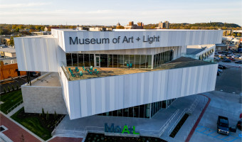 External image of the Museum of Art + Light from roof level featuring walls of windows, an outdoor terrace, and the entrance with "MoA+L" letters.