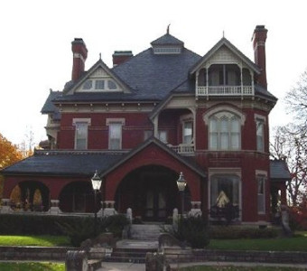 Image of a three storey red brick mansion with an arched entry and large covered wrap porch