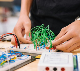 Image of man's hands attaching green wires to a board with various electrical components in the foreground by Jeswin Thomas on Unsplash