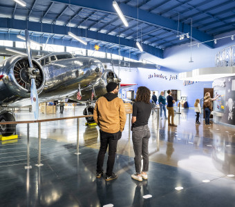 People stand inside the Amelia Earhart Hangar Museum with a silver Muriel Lockheed Electra 10-E airplane behind them.