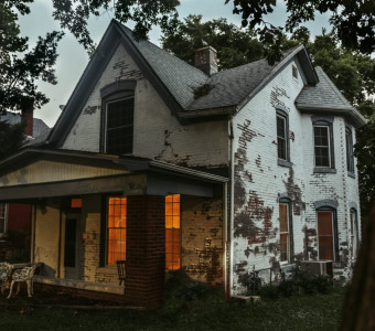 Image of Sallie House in Atchison, a weathered white and red brick two storey house with glowing windows on the first floor under the porch roof.