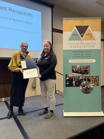 Image of two women standing in front of a projector screen holding a certificate with the KMA stand banner to their right.