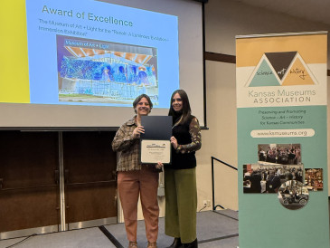 Image of projector screen with Award of Excellence and an image with two women standing in front of it holding a certificate while the KMA stand sign is to their right.