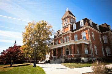 Image of the 3 storey red brick St. Cecilia Hall with tower rising above the 3rd storey, wrap porch, and tree in front