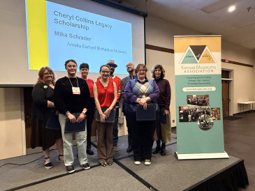 Image of eight KMA Conference Scholarship recipients at the 2025 Conference standing next to KMA's bannerstand with a screen behind displaying the text "Cheryl Collins Legacy Scholarship, Mika Schrader, Amelia Earhart Birthplace Museum"