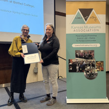 Image of two women standing in front of a projector screen holding a certificate with the KMA stand banner to their right.