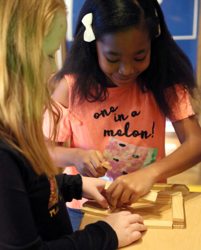 One white girl and one black girl in a shirt that says "one in a melon" with a watermelon on it build with small wooden planks at the Smoky Hill Museum.