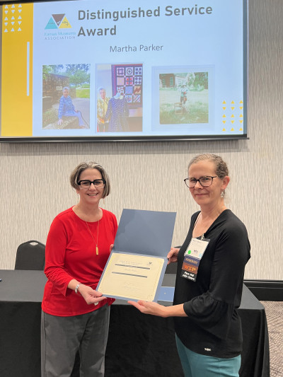 KMA Board Member Dawn Hammatt, left, hands a certificate to Marin Massa, in honor of Martha Parker, recipient of KMA's Distinguished Service Award while images of Martha appear on a screen behind them.