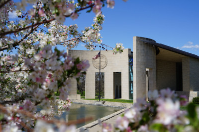 A an exterior photo of the Dole Institute (curved portico with an American flag) and blooming flowers in the foreground.
