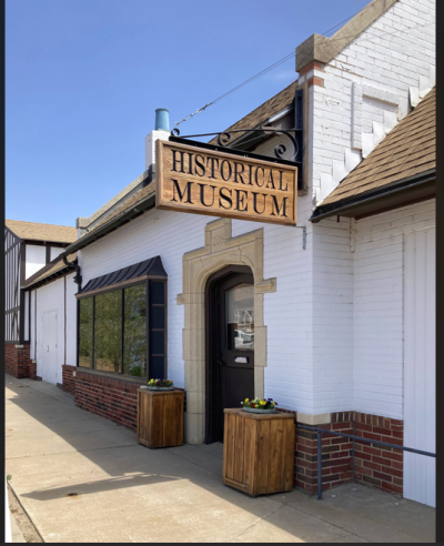 Image of the Pratt Historical Museum, a white painted brick building with 3 bay window and arched stone entrance flanked by wooden planters.  Above hangs a wooden sign that says "Historical Museum"
