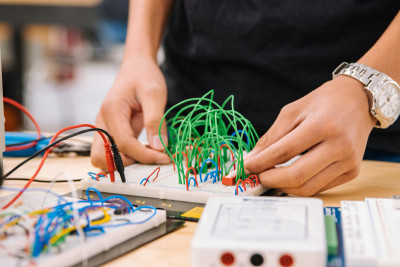 Image of man's hands attaching green wires to a board with various electrical components in the foreground by Jeswin Thomas on Unsplash