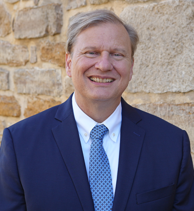 Image of Patrick Zollner, Executive Director of Kansas Historical Society, standing in front of a stone wall wearing a navy blue suit jacket, white collared shirt and light blue patterned tie