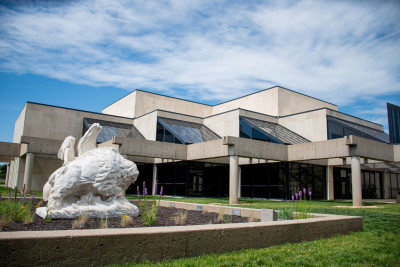 Image of the concrete and glass Kansas Historical Society with a blue sky and clouds above.  A statute of an Indigenous person, horse, and bison are in the left foreground inside a landscaped area.