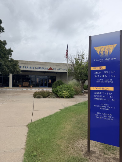 Exterior image of the Prairie Museum of Art and History with a flagpole and blue sign with visitor information in the foreground.