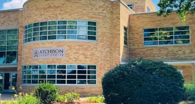 Image of the mottled tan brick and glass Atchison Event Center with the name sign in the middle and bushes in front.