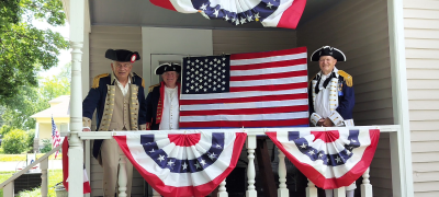 Three men dressed in Revolutionary War uniforms stand on a porch dressed in red white and blue buntings while two of them display an American flag.