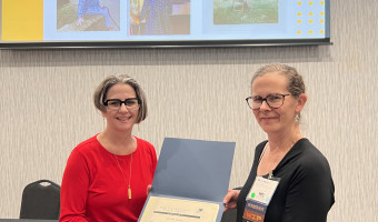 KMA Board Member Dawn Hammatt, left, hands a certificate to Marin Massa, in honor of Martha Parker, recipient of KMA's Distinguished Service Award while images of Martha appear on a screen behind them.