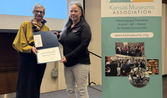 Image of two women standing in front of a projector screen holding a certificate with the KMA stand banner to their right.