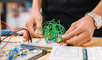 Image of man's hands attaching green wires to a board with various electrical components in the foreground by Jeswin Thomas on Unsplash
