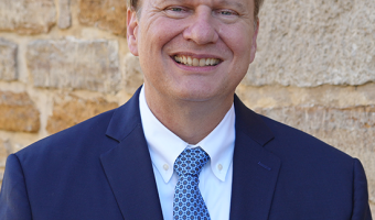 Image of Patrick Zollner, Executive Director of Kansas Historical Society, standing in front of a stone wall wearing a navy blue suit jacket, white collared shirt and light blue patterned tie
