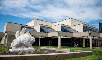 Image of the concrete and glass Kansas Historical Society with a blue sky and clouds above.  A statute of an Indigenous person, horse, and bison are in the left foreground inside a landscaped area.