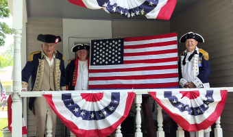 Three men dressed in Revolutionary War uniforms stand on a porch dressed in red white and blue buntings while two of them display an American flag.